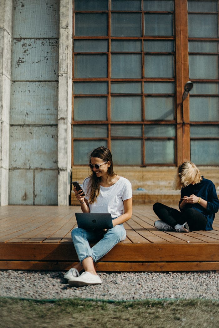 Two women working remotely on laptops outdoors, enjoying the sun. Perfect for freelance lifestyle themes.